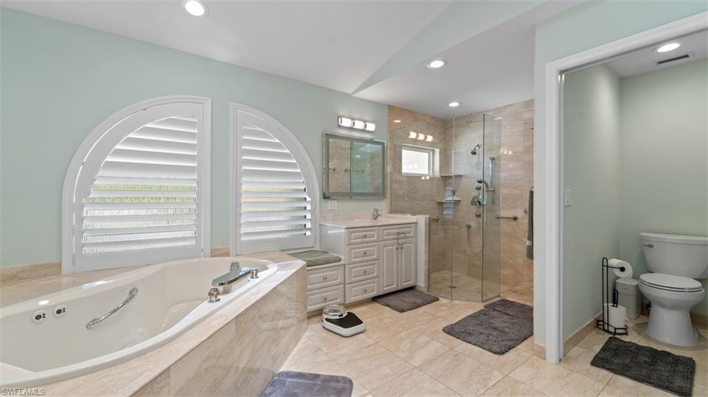 Spa-inspired bathroom featuring a jetted tub, arched plantation shutters, a glass-enclosed shower, a double vanity with a marble countertop, and recessed lighting