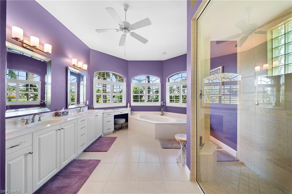Dual vanities with undermount sinks, built-in makeup desk, arched windows with plantation shutters, corner soaking tub, and a glass-enclosed shower with tiled walls