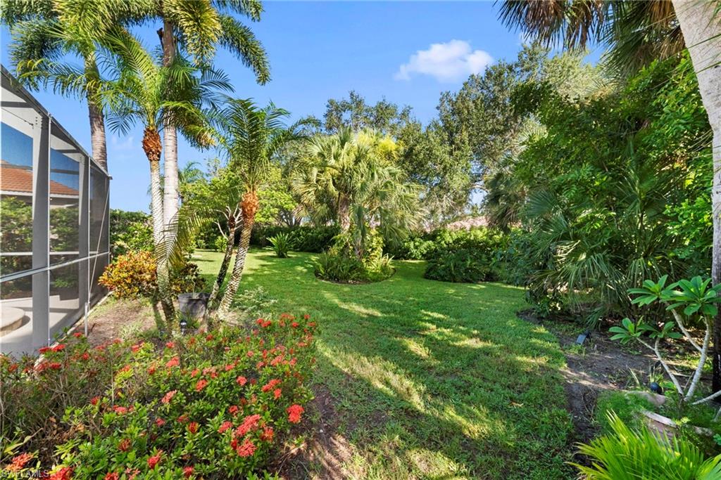 Expansive lawn bordered by mature tropical landscaping, featuring vibrant red flowering bushes and a screened enclosure