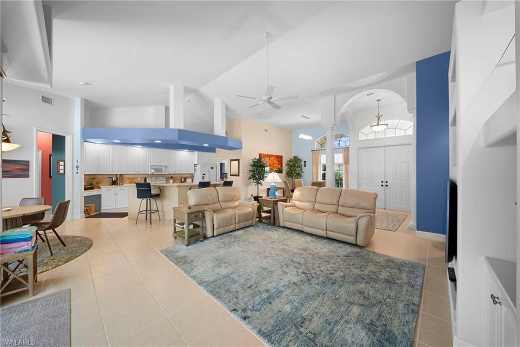 Open-concept living space featuring high vaulted ceilings, light-toned tile flooring, a kitchen with white cabinetry and a built-in breakfast bar, and a double-door entry with an arched transom
