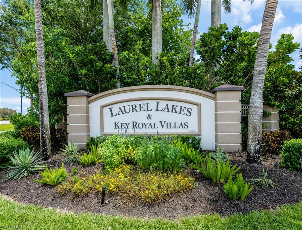 Community entrance sign for Laurel Lakes & Key Royal Villas featuring a stucco and stone finish, surrounded by varied tropical landscaping, mature palm trees, and manicured shrubbery