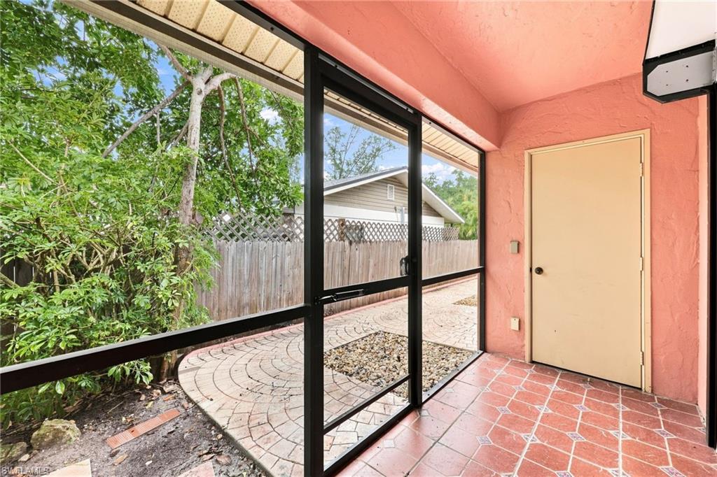 Covered lanai with black-framed glass panels and coral terracotta tile flooring, overlooking circular-patterned paver hardscaping and mature landscaping