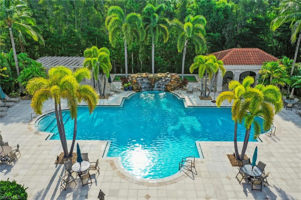 Expansive outdoor pool area featuring a custom-shaped swimming pool with a rock waterfall feature, surrounding paver patio, a covered cabana with a tile roof, and extensive tropical landscaping