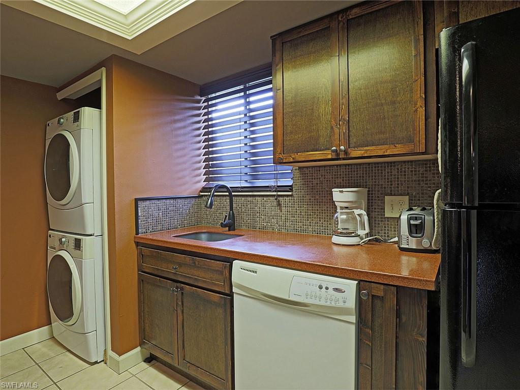Compact kitchen area featuring a stacked washer and dryer, dark wood-finish cabinetry, a white dishwasher, a black refrigerator, and a tiled backsplash