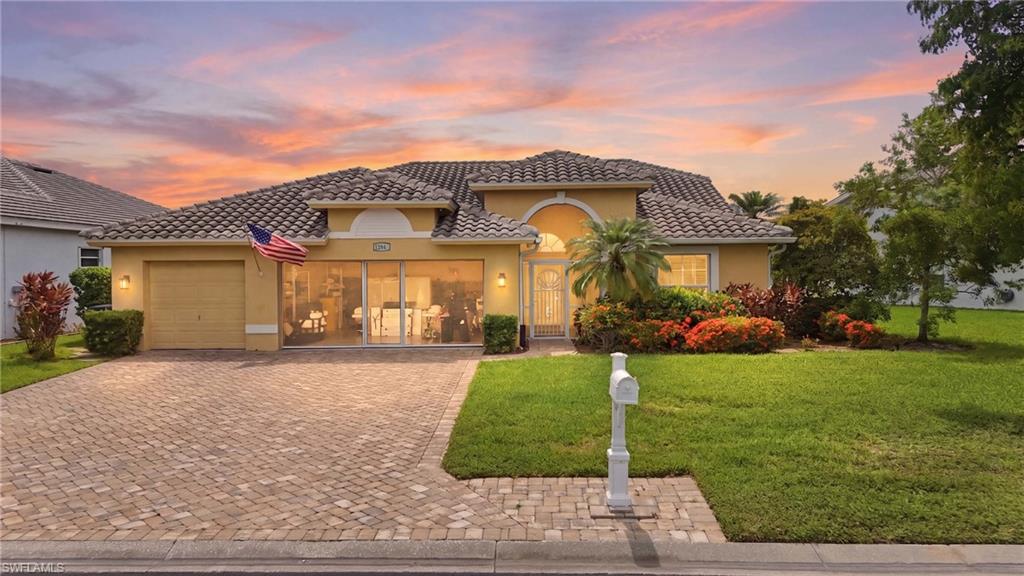 Single-story stucco residence featuring a tile roof, paver driveway, an arched entry, and extensive front landscaping with mature shrubs and trees