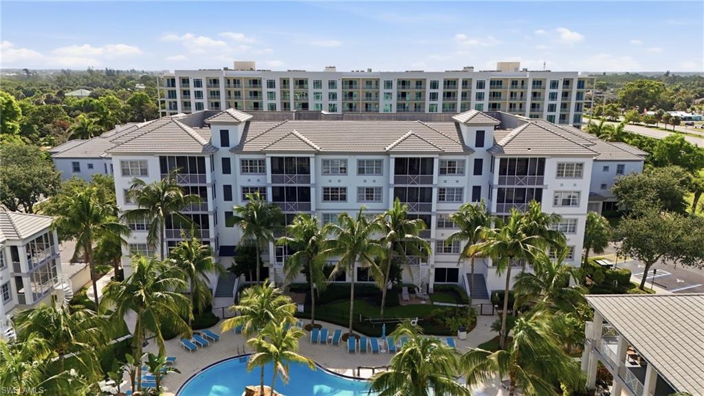 Multi-story residential complex featuring screened balconies, a central swimming pool with lounge chairs, mature palm trees, and a light-colored exterior with gray roofs