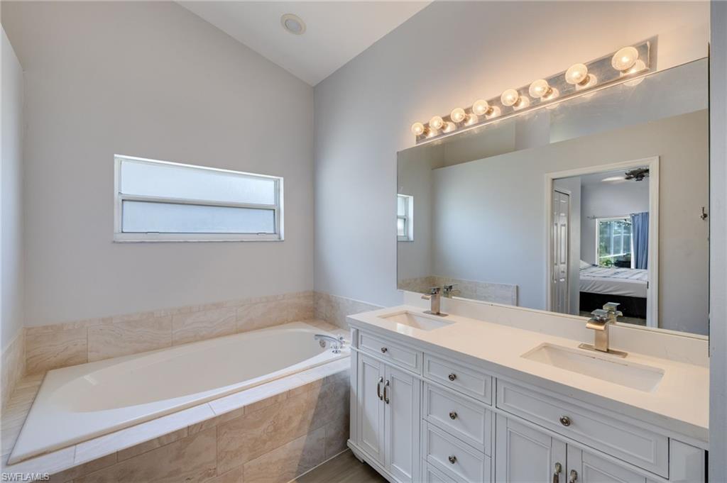 Bathroom featuring a dual vanity with white cabinetry and integrated sinks, a large mirror with strip lighting, a built-in bathtub with tiled surround, and a clerestory window
