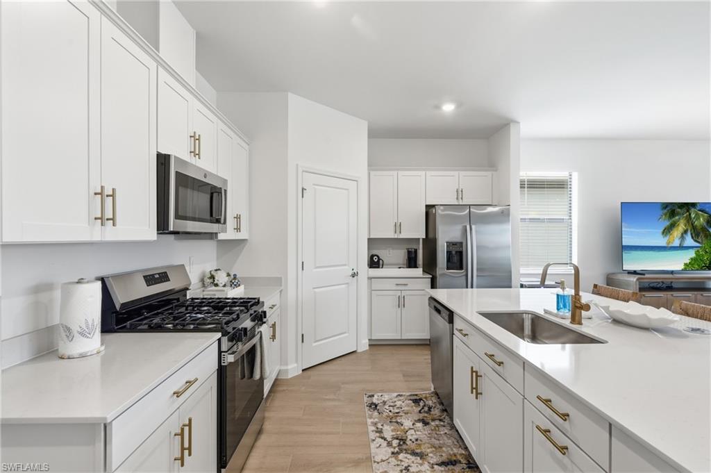 Modern kitchen featuring white cabinetry with gold-tone hardware, stainless steel appliances, light-toned countertops, and wood-finish flooring