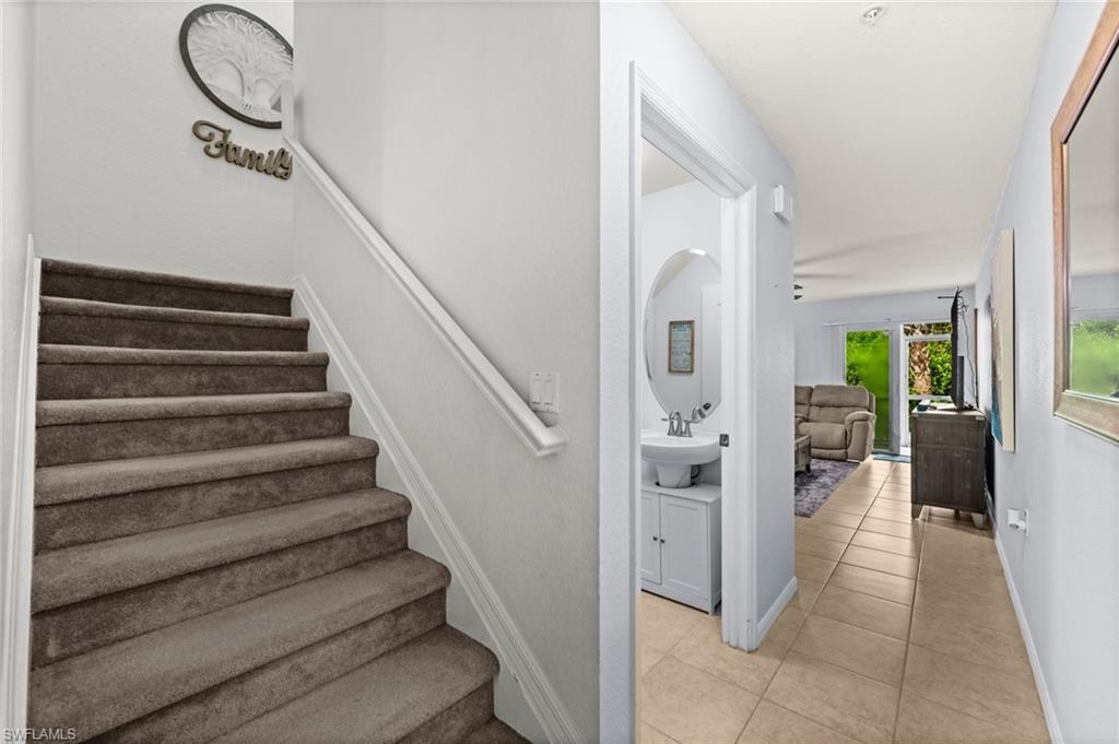 Tiled hallway entry featuring a half bath with an oval mirror and a white vanity with a basin sink, leading to a living area with a glass sliding door and a carpeted staircase with a white handrail