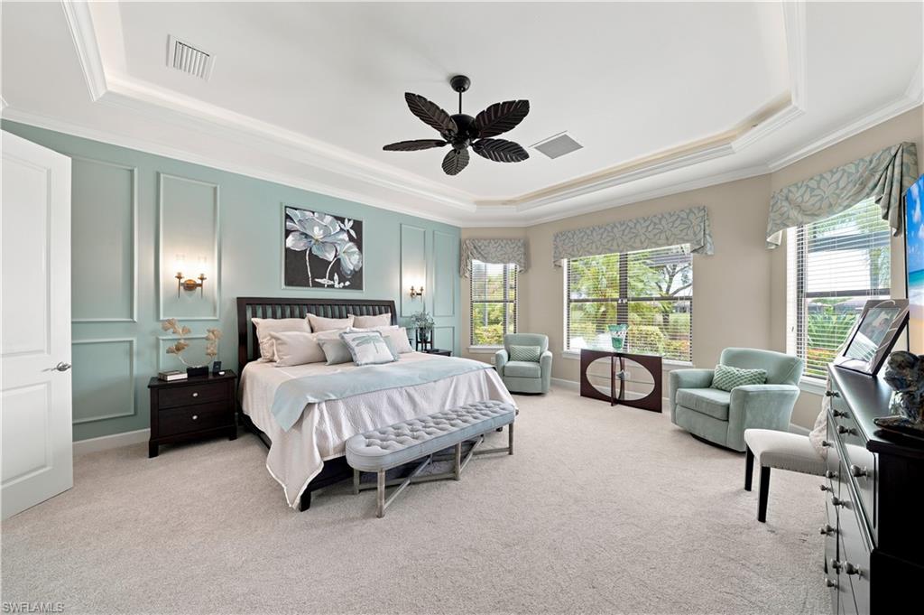 Spacious bedroom featuring light-colored carpeting, a tray ceiling with crown molding, and a dark-toned ceiling fan