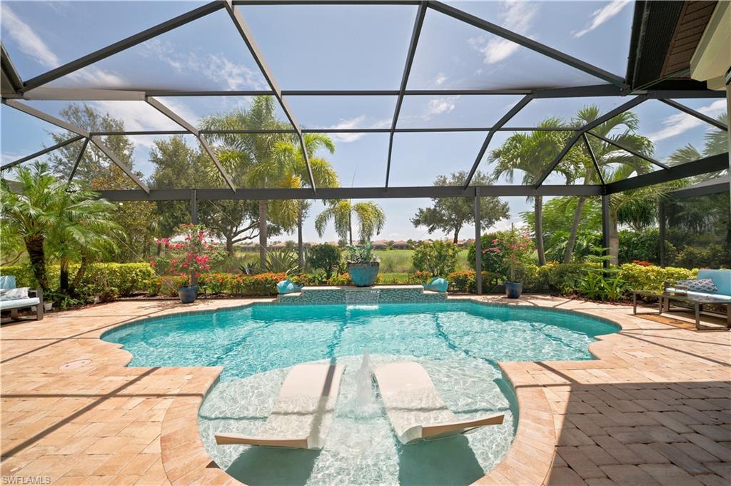 Screened pool enclosure featuring a freeform pool with sun shelf and water feature, surrounded by paver decking and lush landscaping
