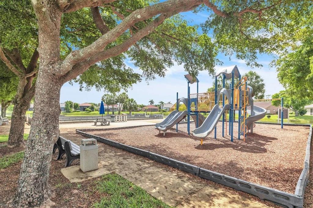 Outdoor recreational area featuring a multi-slide play structure, wood chip ground cover, mature trees providing shade, a black slatted bench, and a nearby picnic table with an umbrella