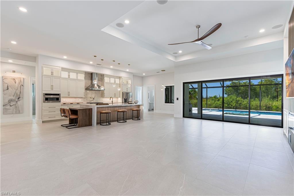 Spacious open-concept living area featuring a gourmet kitchen, large format tile flooring, a tray ceiling with recessed lighting and a ceiling fan, and expansive sliding glass doors opening to an outdoor screened enclosure with a pool
