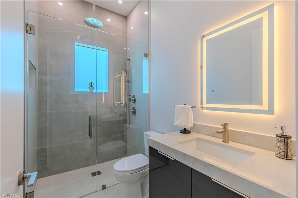 Modern bathroom featuring a frameless glass shower enclosure with a rainfall showerhead, a large vanity with an integrated sink and light-colored countertop, dark cabinetry with bar pulls, and a backlit mirror