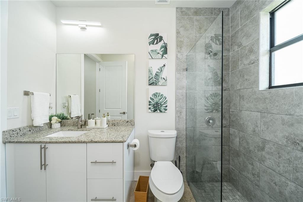 Bathroom featuring a frameless glass shower enclosure, stone-look tile walls, a vanity with a light-colored countertop, and a white toilet