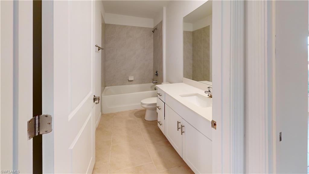 Bathroom featuring a white vanity with integrated sink, a rectangular mirror, a bathtub with tiled surround, a toilet, and light-toned floor tiling