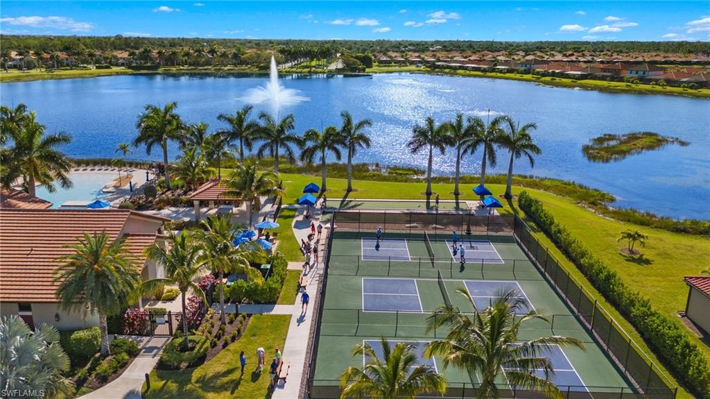 Expansive community amenities featuring multiple pickleball courts, a large lake with fountain, mature palm trees, lush landscaping, and a clubhouse with a terracotta tile roof