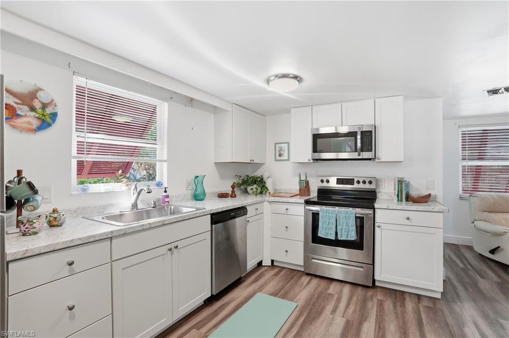 Kitchen featuring white shaker-style cabinetry, stainless steel appliances, a double basin sink, light-toned countertops, and wood-finish flooring - Virtually Edited Image