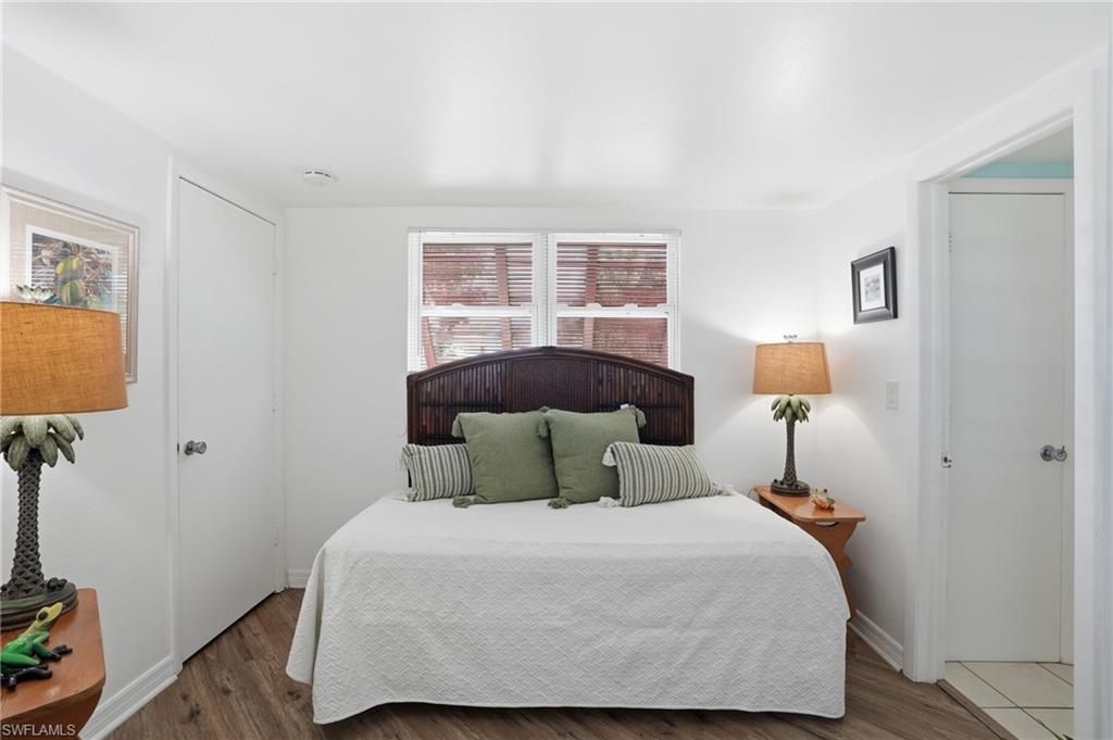 Bedroom featuring wood-finish flooring, a window with blinds, a white door, and a second white door opening to a tiled area - Virtually Edited Image