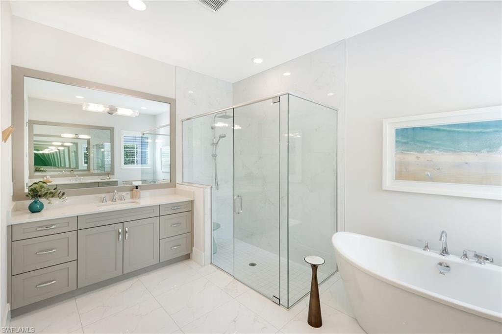 Spacious bathroom featuring a frameless glass shower enclosure, a freestanding soaking tub, dual vanity with light gray cabinetry, a large framed mirror, and white tile flooring
