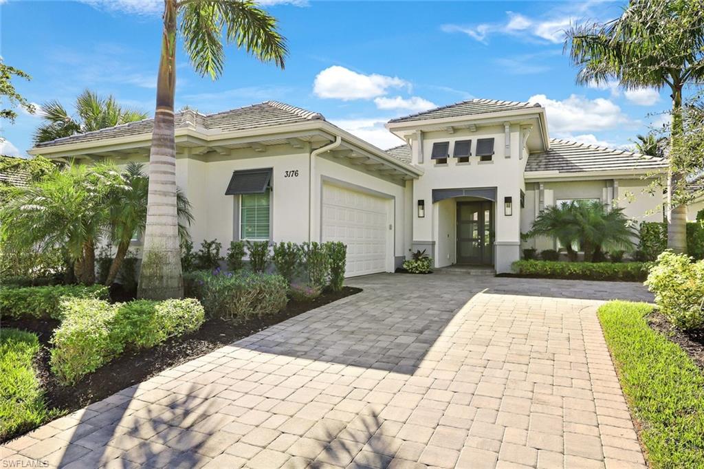 Contemporary residence featuring a paver driveway, an attached garage, a tile roof, a covered entry with dual sconces, and mature tropical landscaping