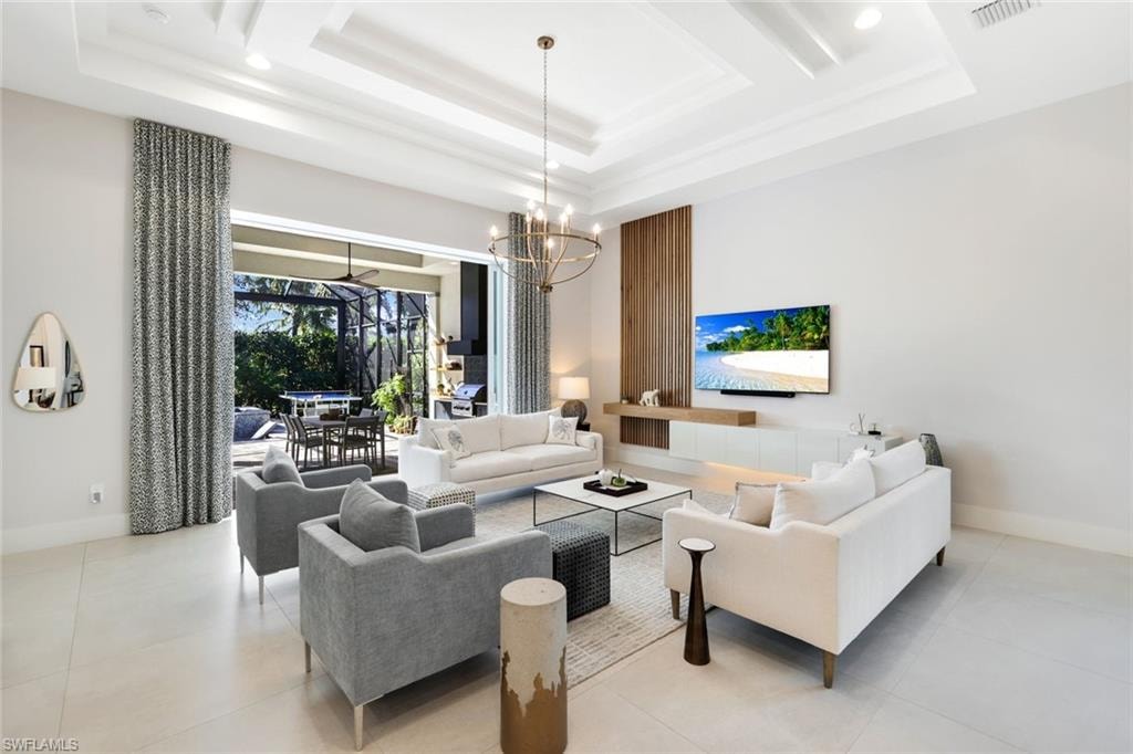 Spacious living area featuring high ceilings with tray detailing and recessed lighting, large format tile flooring, a custom accent wall with integrated wood-finish paneling and floating cabinetry, and expansive sliding glass doors opening to an outdoor living space with a screened enclosure