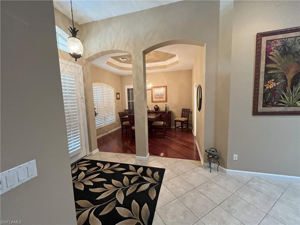 Tiled entryway with an arched doorway opening to a dining space featuring wood-finish flooring, plantation shutters, and a tray ceiling
