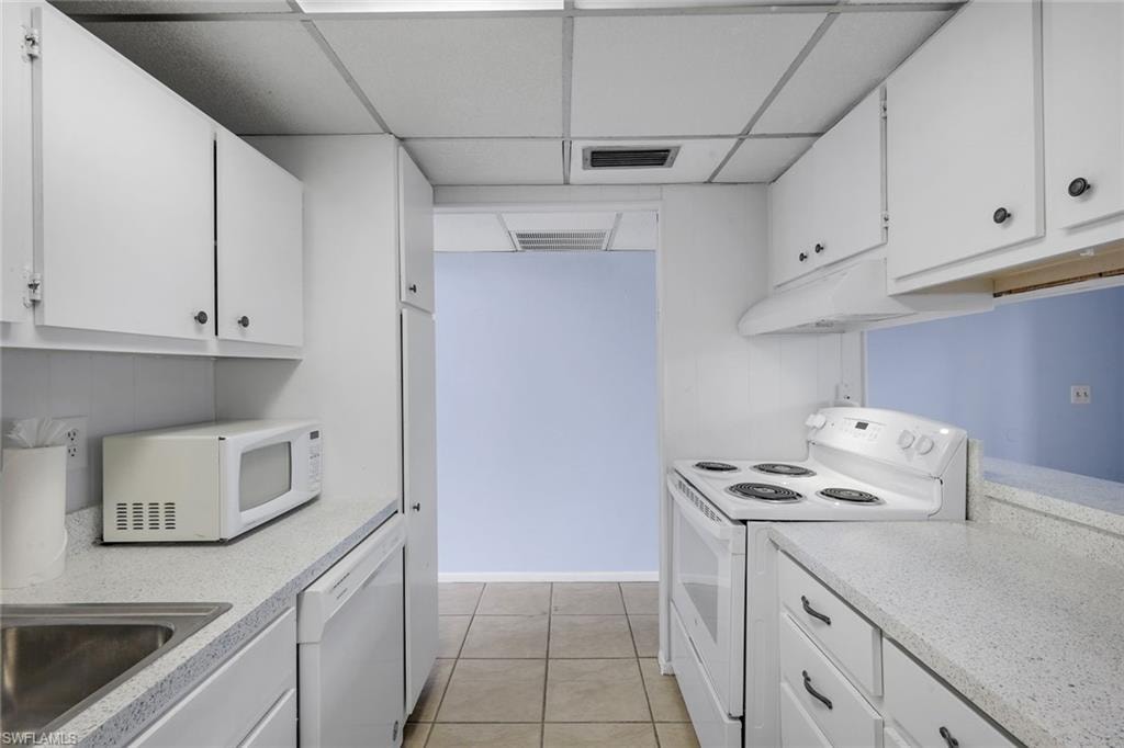 Galley kitchen featuring white cabinetry, light speckled countertops, a double basin stainless steel sink, tile flooring, and white appliances including a range and dishwasher