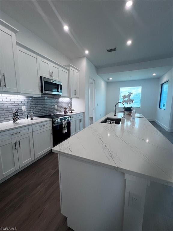 Contemporary kitchen featuring an oversized island with a built-in sink and dark pull-down faucet, white shaker cabinetry, stainless steel appliances, and wood-finish flooring