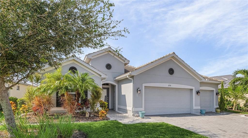 Gray stucco exterior with a tile roof, featuring a paver driveway, an integrated two-car garage, and a lush tropical landscape with mature trees