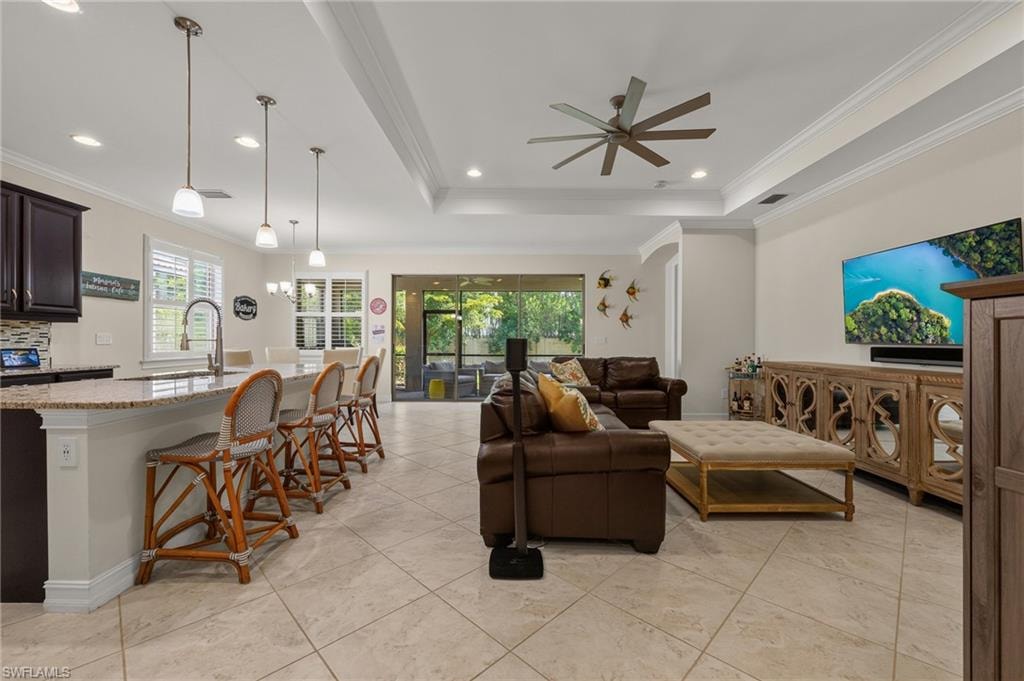 Open-concept living space featuring neutral tile flooring, tray ceilings with crown molding, recessed lighting, a modern ceiling fan, and a kitchen island with pendant lighting