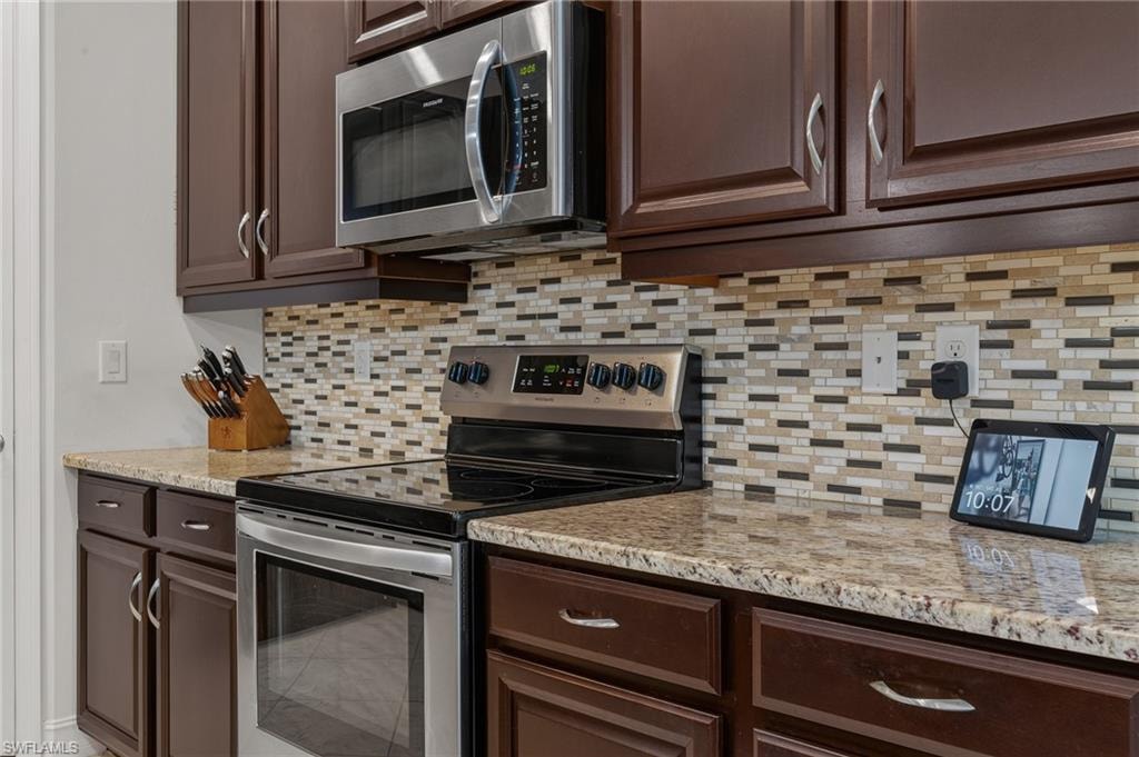 Kitchen featuring dark wood cabinetry with chrome pulls, stainless steel appliances, a mosaic tile backsplash, and light-toned granite countertops