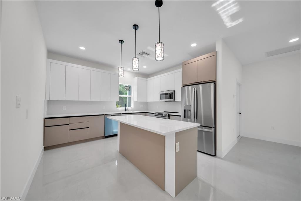 Modern kitchen featuring a central island with a solid surface countertop, stainless steel appliances, white flat-panel upper cabinetry, taupe flat-panel lower cabinetry, and recessed lighting