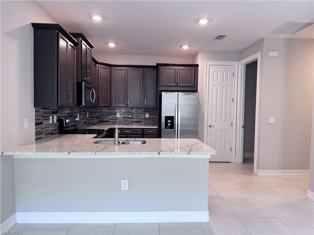 Kitchen with backsplash, appliances with stainless steel finishes, dark brown cabinets, light tile patterned floors, and light stone counters