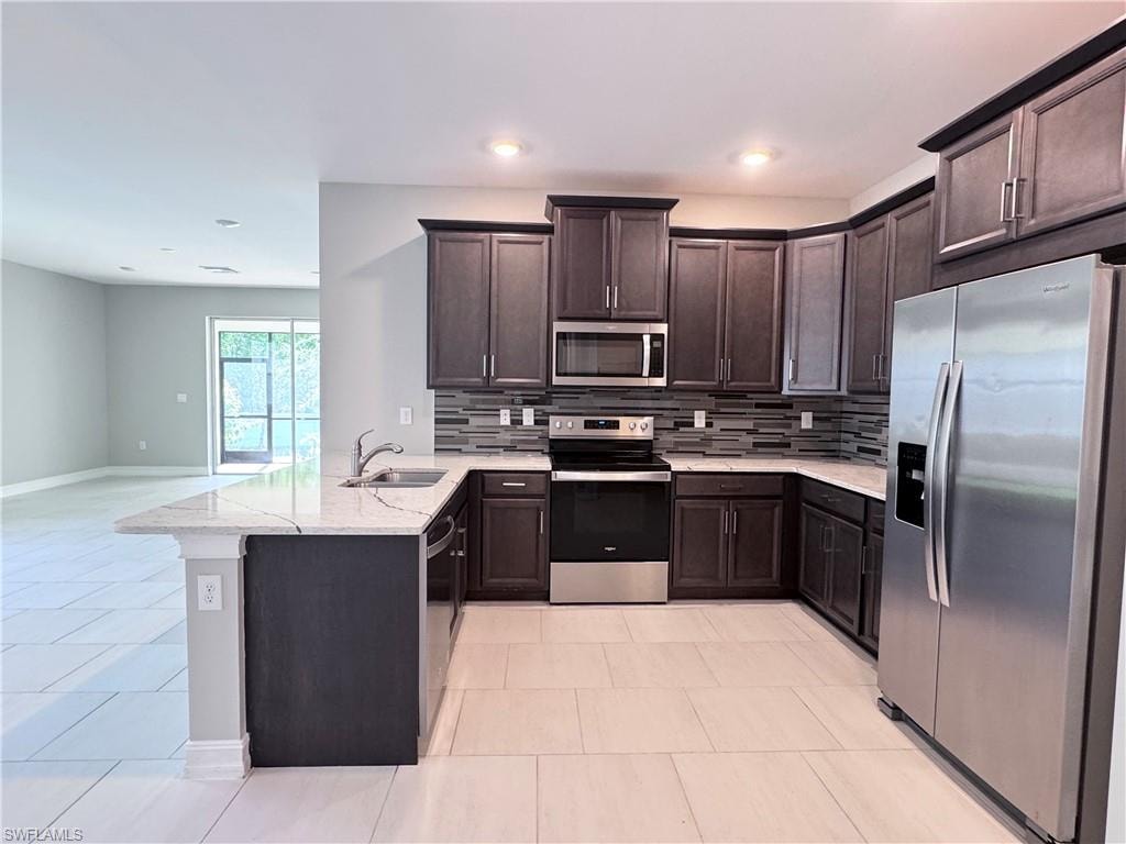 Kitchen with stainless steel appliances, dark brown cabinetry, a peninsula, light stone counters, and decorative backsplash