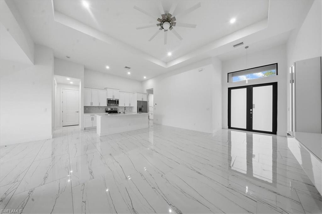 Spacious open-concept interior featuring large format marble-look tile flooring, a contemporary kitchen with white cabinetry and stainless steel appliances, a tray ceiling with recessed lighting, and a modern ceiling fan