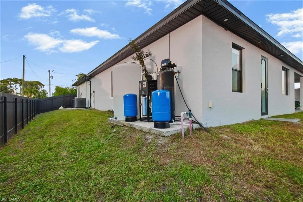 White stucco exterior with a dark fascia and soffit, featuring a black privacy fence, a visible HVAC unit, and an exterior water treatment system on a concrete pad