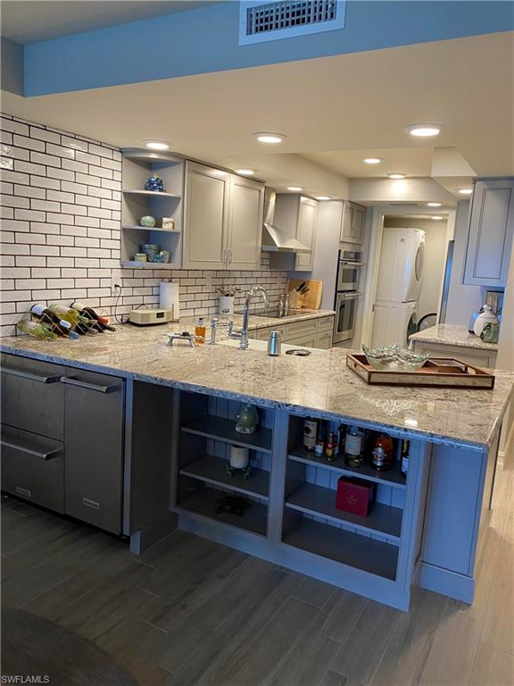 Kitchen featuring light stone countertops, dark hardwood / wood-style flooring, kitchen peninsula, wall chimney range hood, and double oven