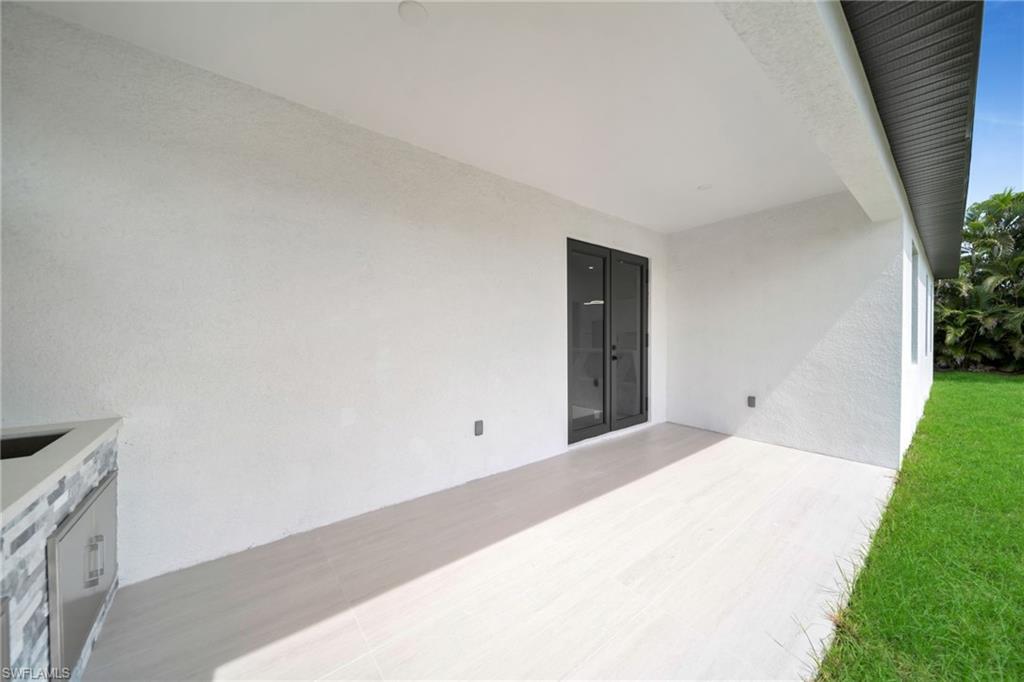 Covered patio with a light-colored flooring, featuring double glass doors, a built-in outdoor counter with a stainless steel cabinet, and a textured white stucco exterior