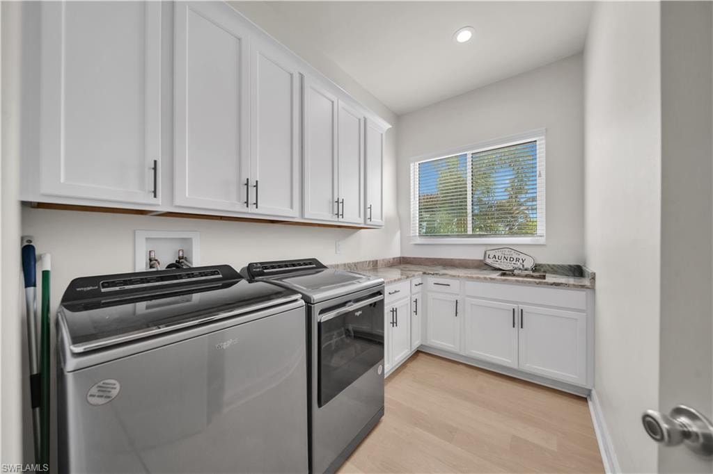 Modern laundry room featuring stainless steel front-load washer and dryer with a sleek gray finish, white shaker-style cabinetry with brushed nickel hardware, quartz countertops, and abundant natural light from a large dual-pane window