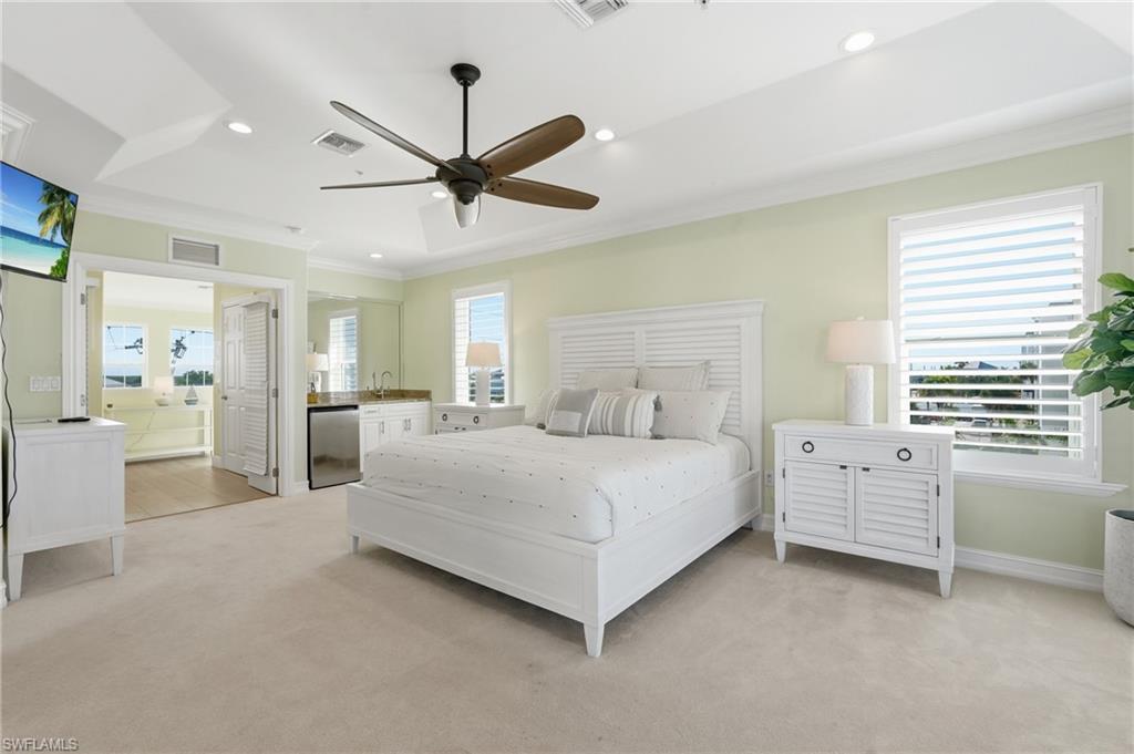 Tray ceiling with recessed lighting, ceiling fan, built-in wet bar with stainless steel under-counter refrigerator, two windows with plantation shutters, and light-tone carpeting - Virtually Edited Image