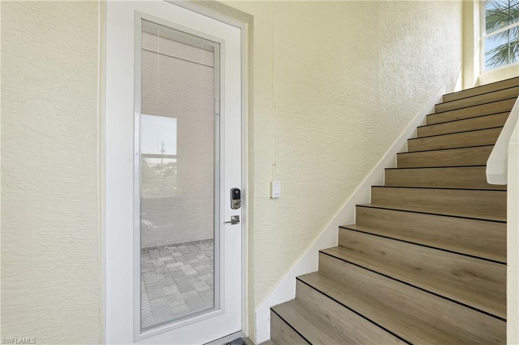 Full-lite glass door with a built-in smart lock, wood-finish staircase with white risers, textured wall finish, and upper-level window providing natural illumination - Virtually Edited Image