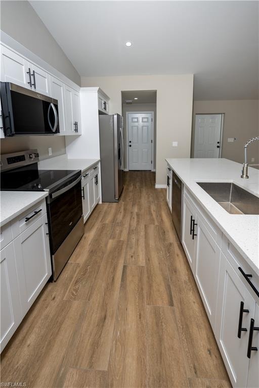 Wood-finish flooring, white cabinetry with black hardware, stainless steel appliances, white countertops, and a kitchen island with an integrated sink