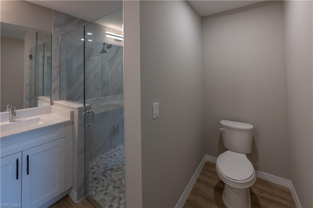White vanity with integrated sink, glass-enclosed shower, patterned shower floor, wood-finish flooring, and a commode