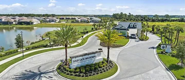 Gated community entrance featuring a prominent stone monument, lush tropical landscaping with palm trees, a grand entrance building with a metal roof, and a large body of water bordering the community