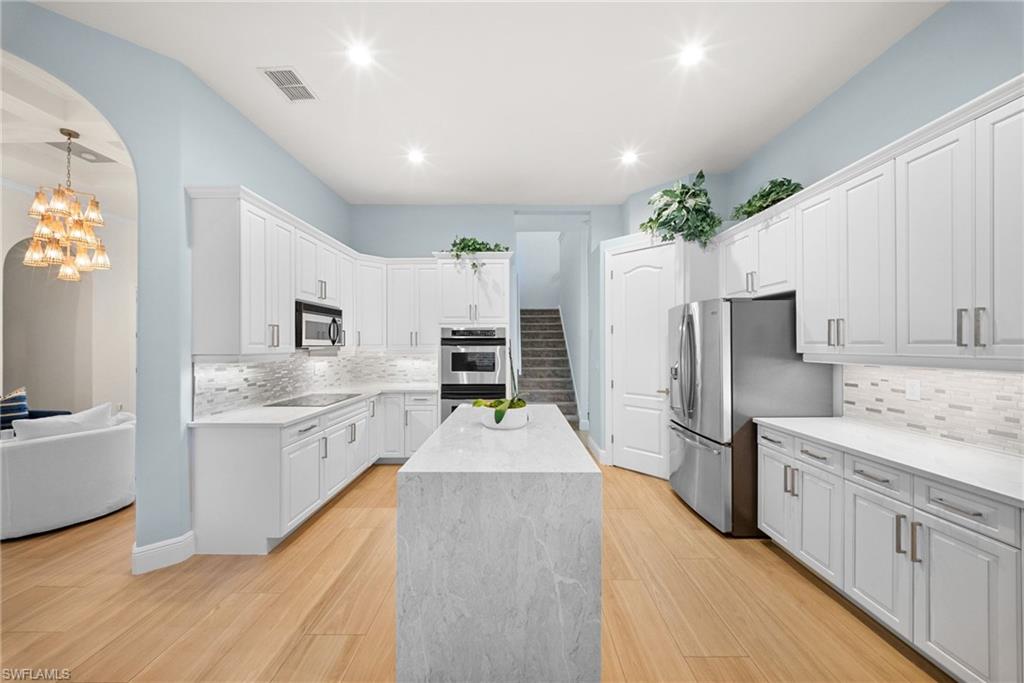 Modern kitchen featuring a central island with a waterfall edge, extensive white cabinetry, stainless steel appliances, light wood-finish flooring, and a tile backsplash