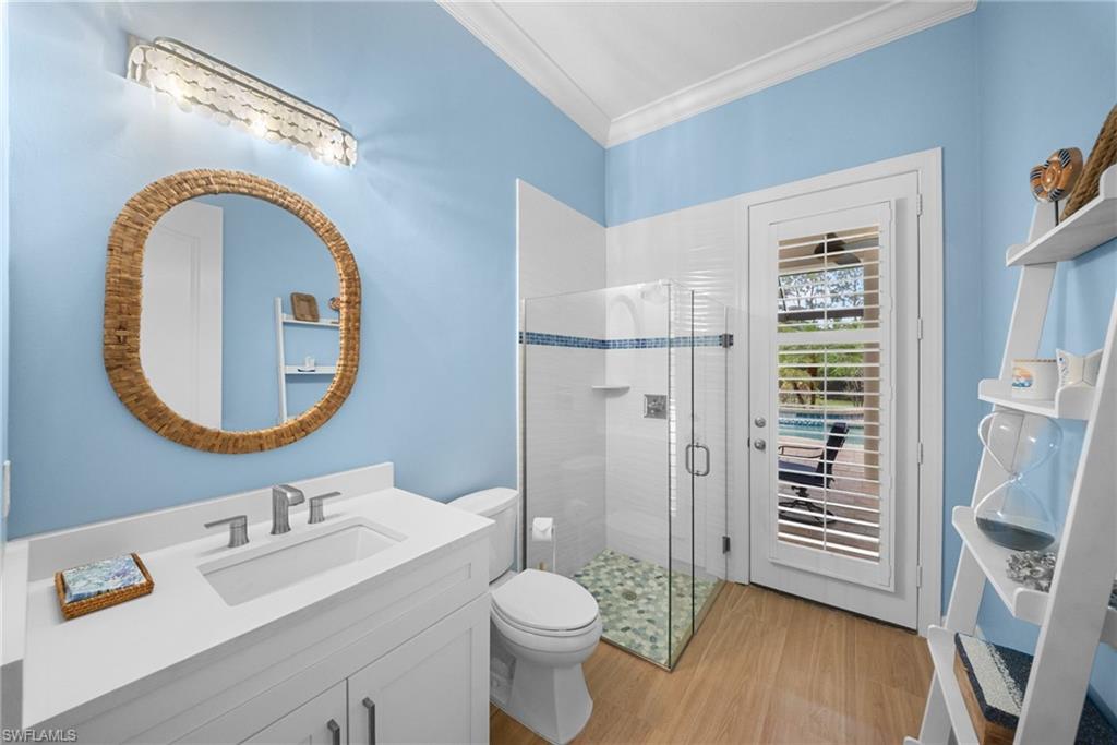 Pool bathroom featuring a white vanity with an integrated sink, a glass-enclosed shower with a pebble floor, a white toilet, and wood-finish flooring, exterior door, accessible by pool area