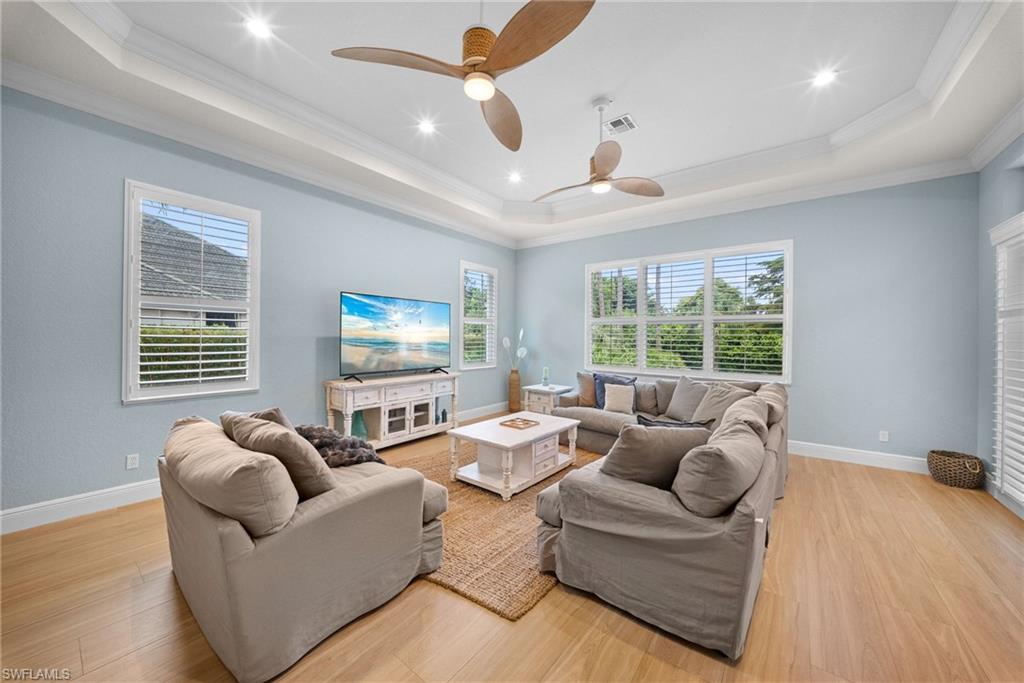 Living area featuring light wood-finish flooring, multiple windows with plantation shutters, tray ceiling with recessed lighting, and two ceiling fans