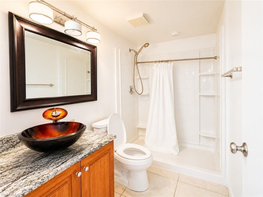 Bathroom featuring a vessel sink with a unique faucet, granite-finish countertop, wood-finish vanity, large framed mirror, and a shower with built-in shelving