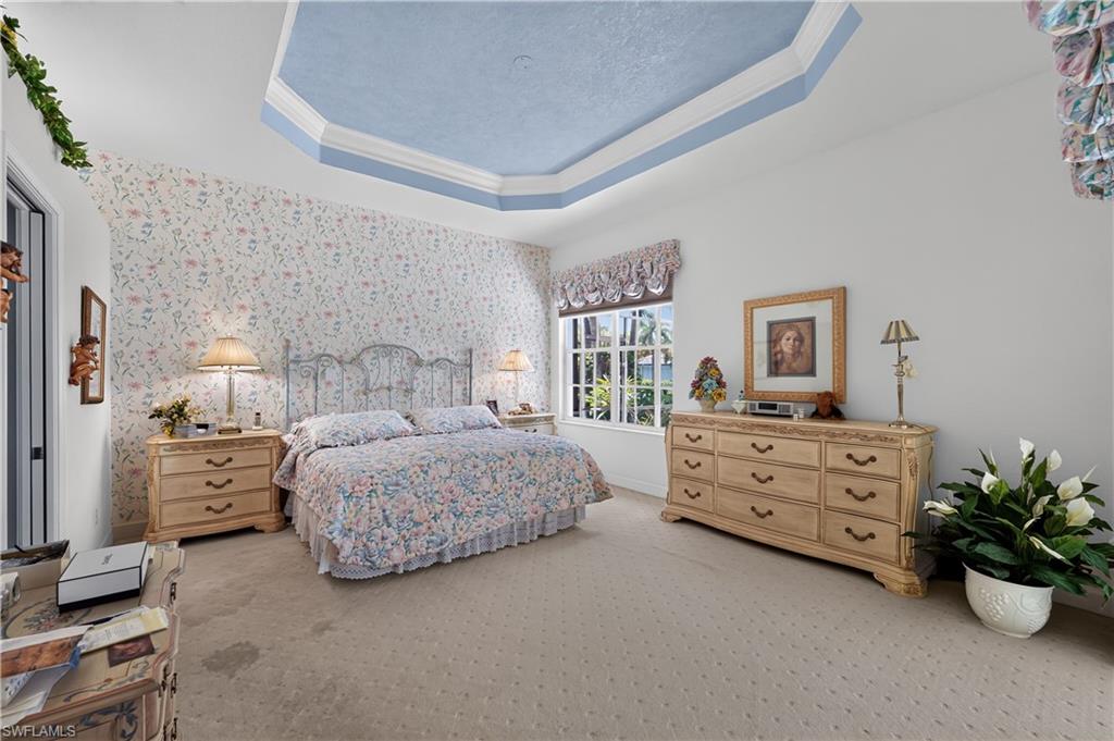Recessed tray ceiling with light blue accent, wall-to-wall carpeting, window with exterior landscaping visible, and a wall of patterned wallpaper